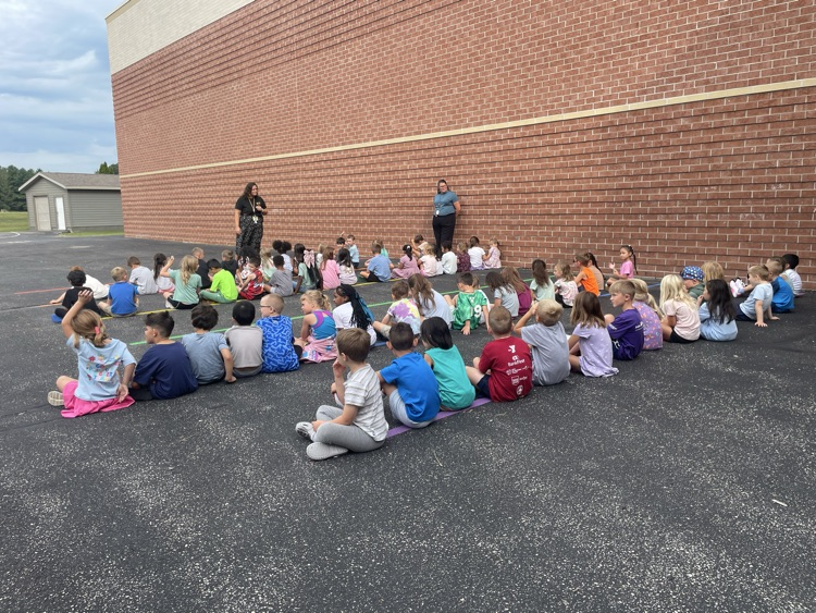 students sitting in rows outside listening to a teacher. 