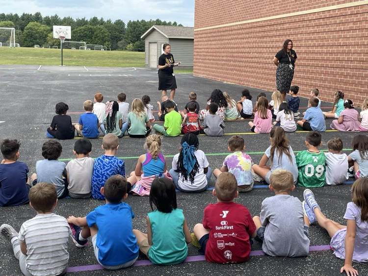 students sitting in rows outside listening to a teacher. 