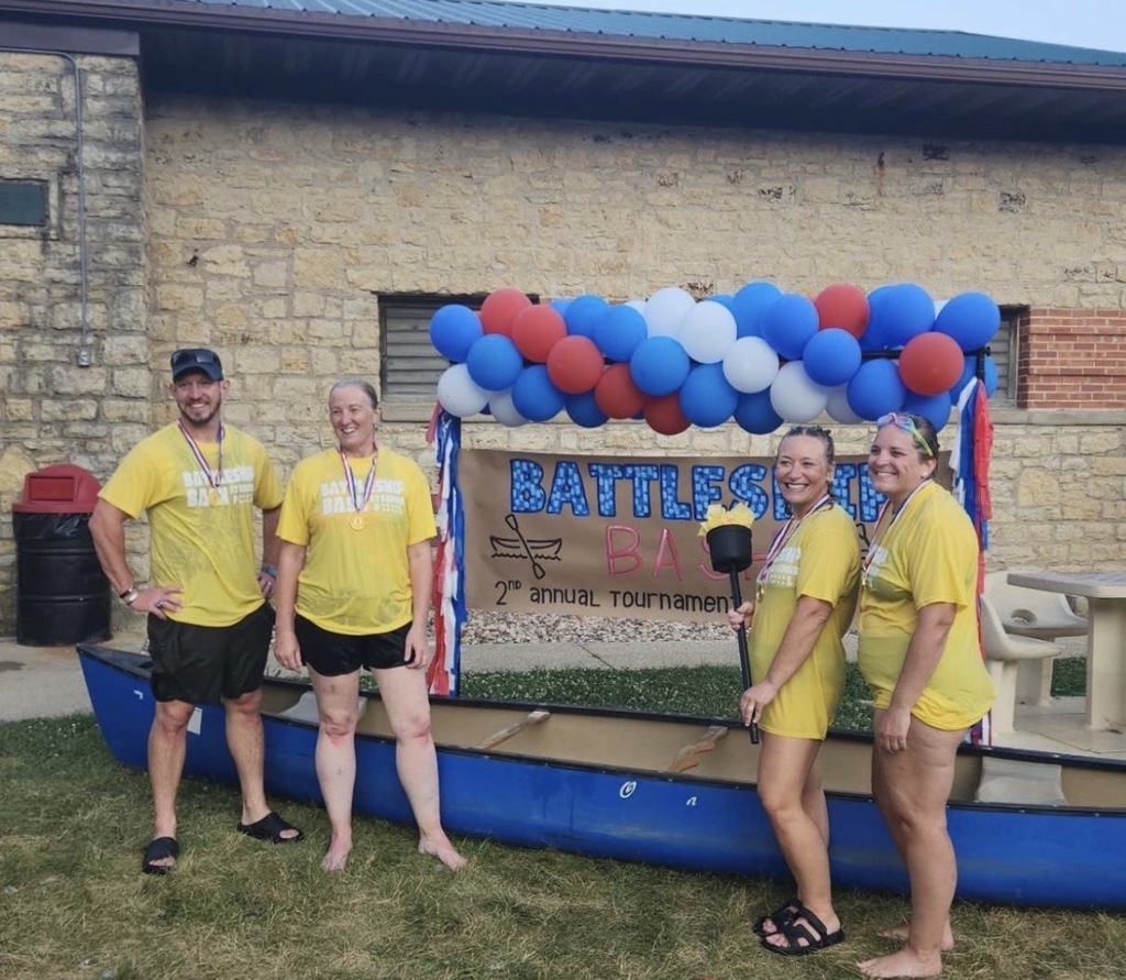 Four people in yellow shirts and shorts pose in front of a boat and a building.