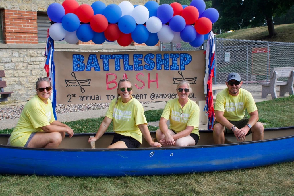 Four people in yellow shirts sit in a canoe with a banner reading "Battleship Bash." Behind them, balloons and a building.