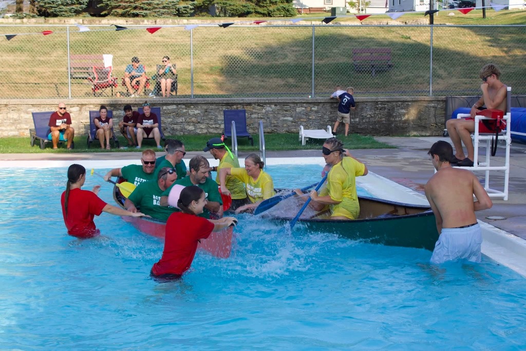 A group of people wearing green and red shirts are rowing a boat in a swimming pool.