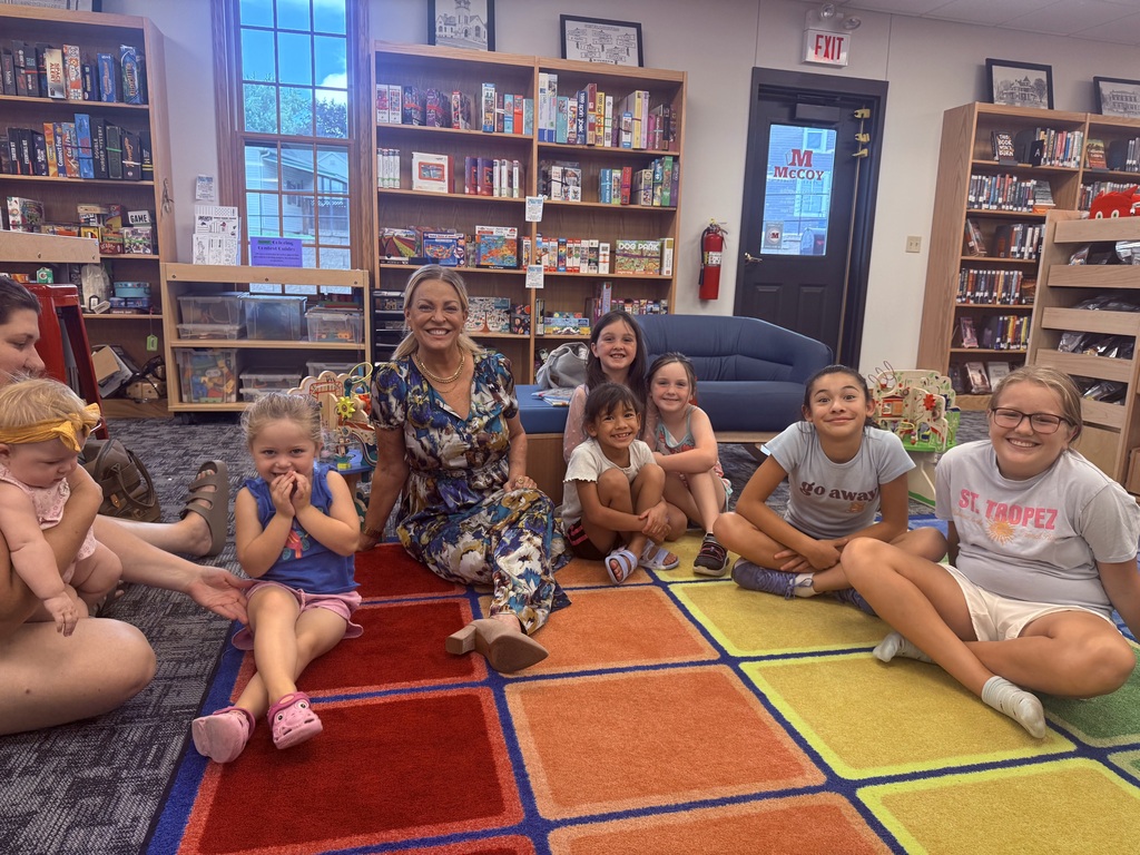 A group of children and adults sit on a colorful rug in a room with bookshelves.