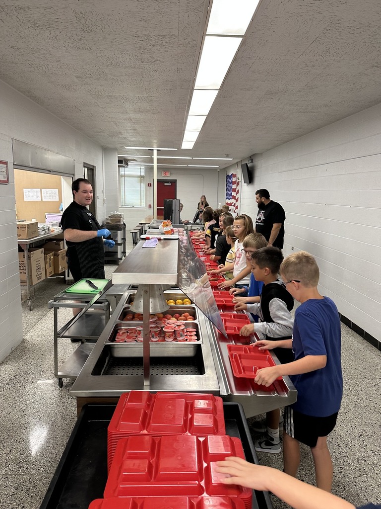Children line up in a school cafeteria to get food served by an adult. Bright lights illuminate the room.