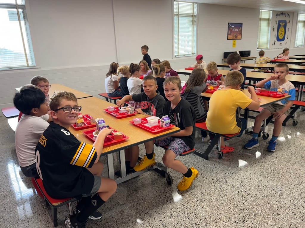 A lunchroom with students eating lunch at tables with red trays. Some students wear sports jerseys and glasses.