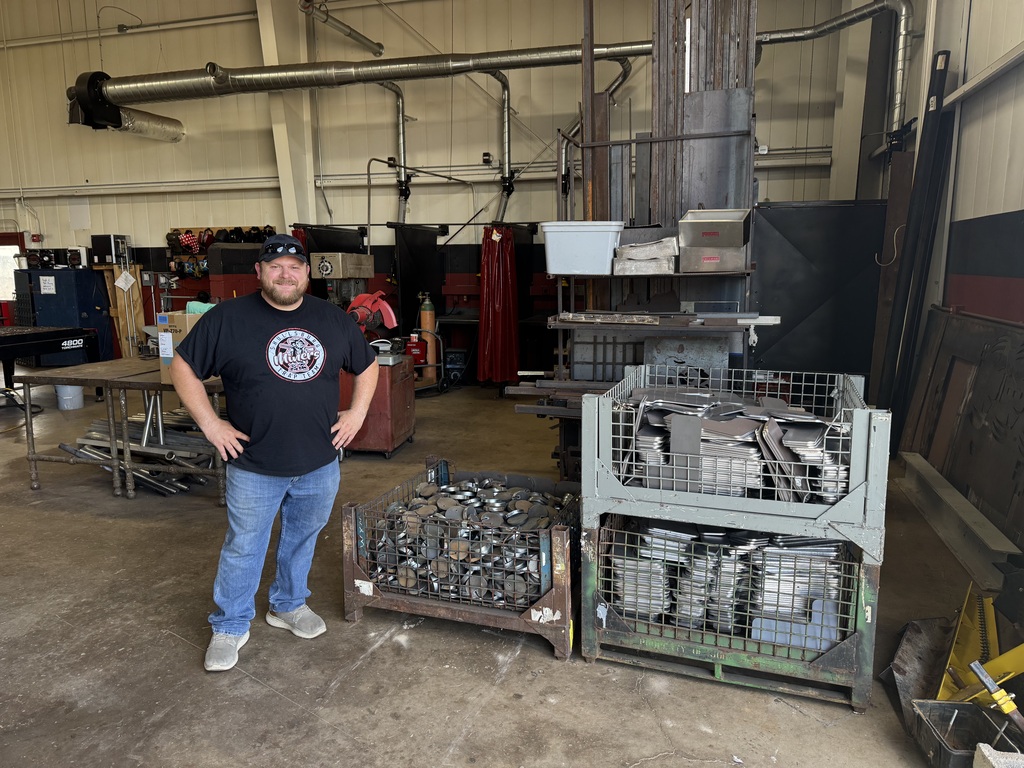 A man stands in a workshop, with metal objects and machinery in the background. He wears a black shirt and jeans.