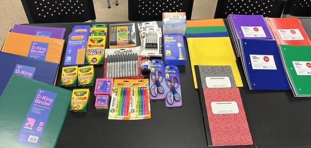 A table displays assorted colorful school supplies, including notebooks, pencils, markers, and more. The items are on a black surface.
