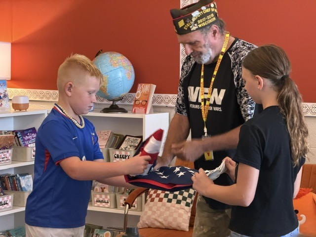 A man with a hat gives a folded flag to a boy and girl in a room.