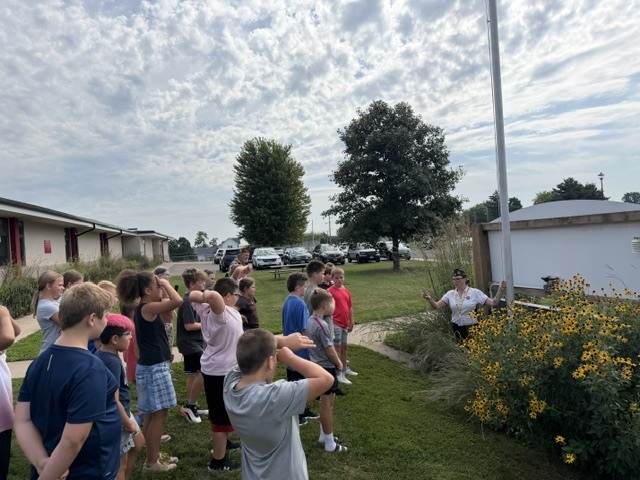 A group of people gather near a building and flagpole, with one person pointing at something.