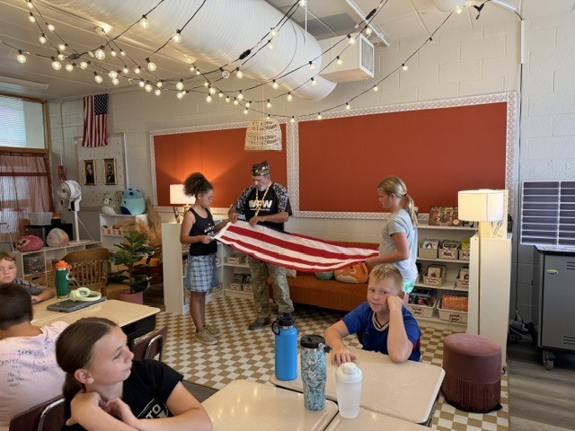 A classroom with students seated. Some students hold an American flag. Red walls and white flooring. Lights hang from the ceiling.