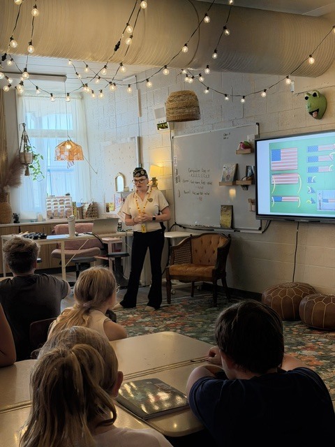 A person in uniform stands before a classroom with students seated. A monitor displays information on the wall.