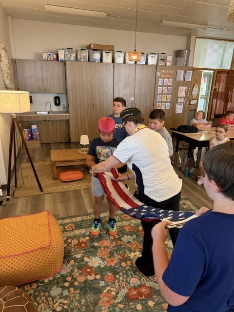 Several children gather around an adult holding an American flag, inside a room with a carpet, wooden floor, and cabinets.