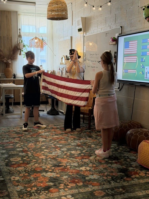 Children holding a flag stand before a screen. One child wears a black shirt, another a pink skirt.