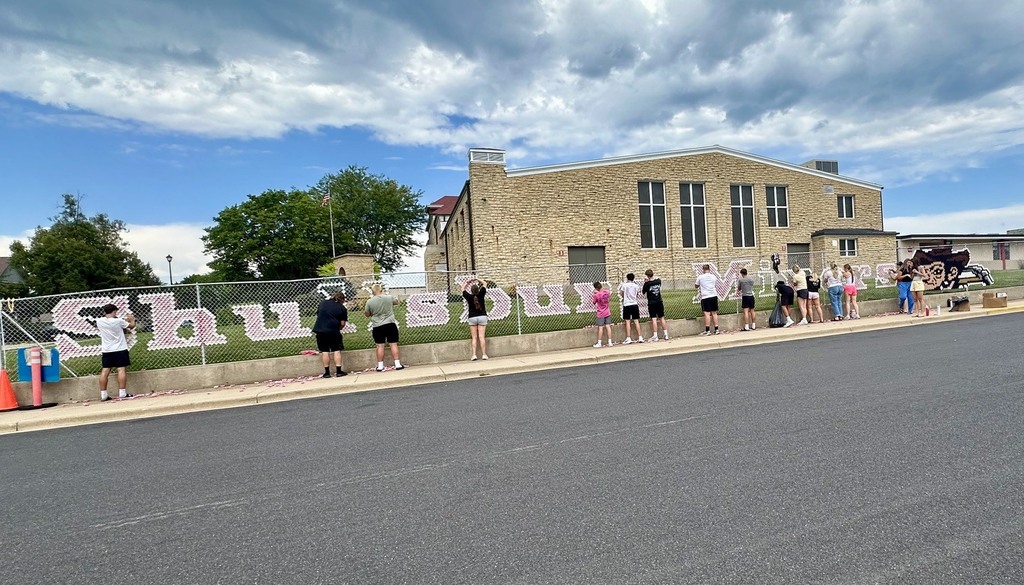 People gather in front of a building with words on the fence.