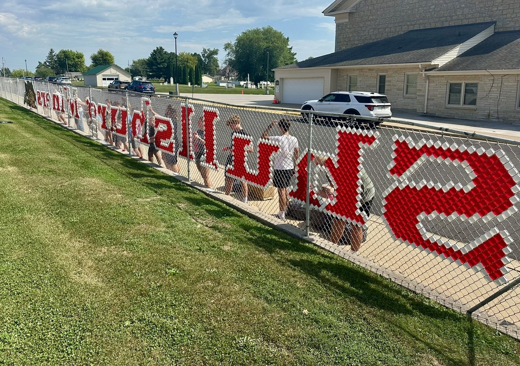 A fence with a red word on it. People stand near it. A building and cars are in the background.