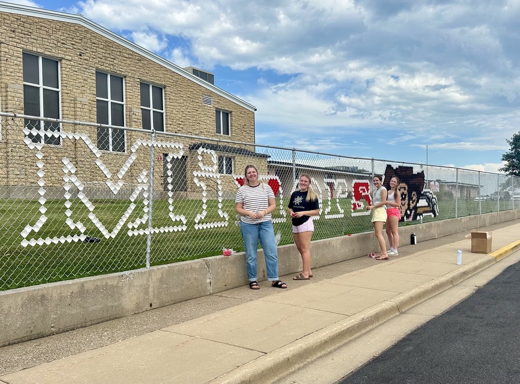 People stand by a fence with large letters, near a building and sidewalk with a grassy area.