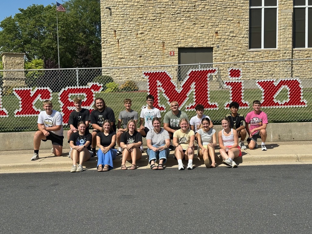 A group of individuals sitting and kneeling outside on a sidewalk, in front of a building with a large sign.