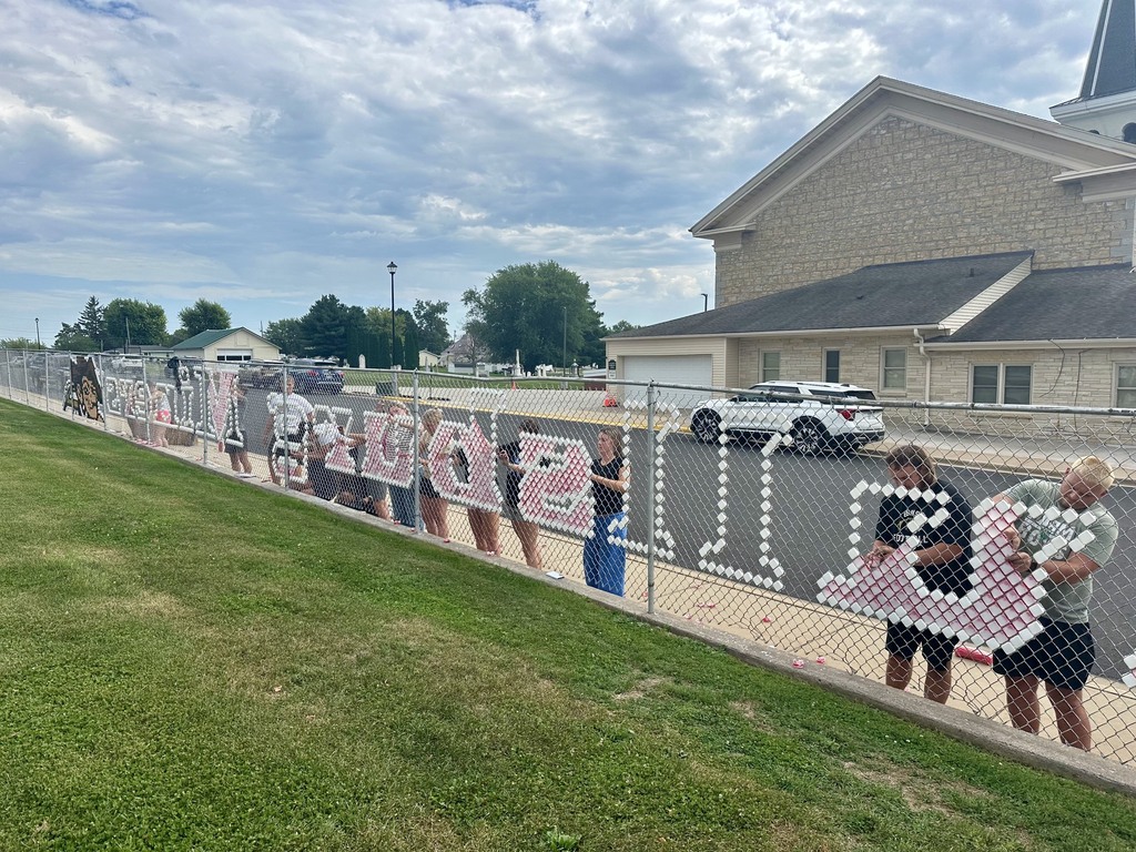 People stand behind a fence with a message. A building is behind them. A vehicle is parked by the driveway.