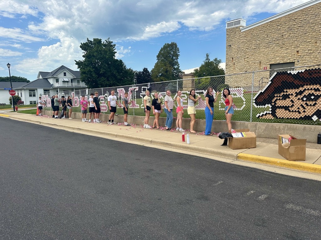 People stand in line outdoors on a sidewalk with a mural behind a fence. A building and houses are in the background.