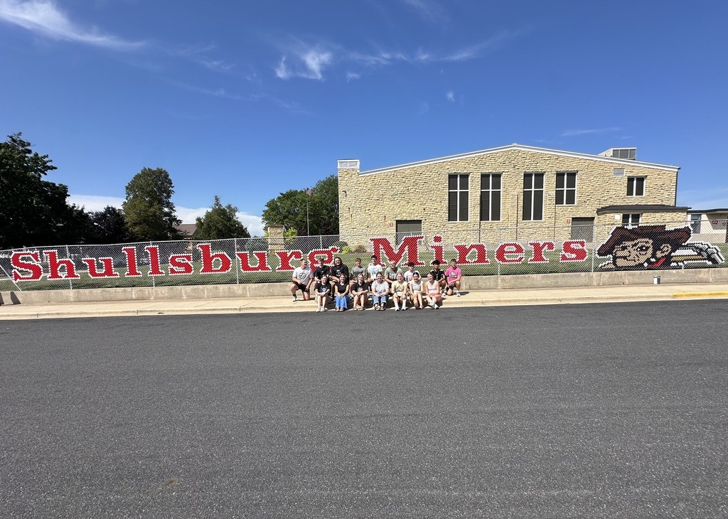 A group of people stand in front of a building with "Shullisburg Miners" painted on the Fence.