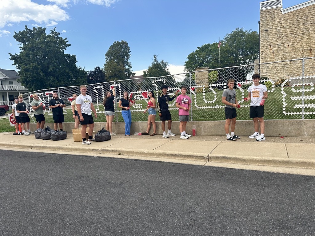 A group of people stands on a sidewalk next to a fence. Letters spell out a word on the fence.