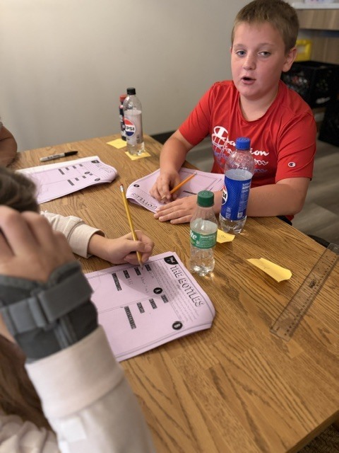 Kids seated at a table. One holds a pencil, focused on a worksheet. A ruler, pens, and water bottles are on the table.