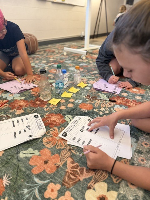 Children sit on a floral blanket and work on papers, with pens, bottles, and notes around them.