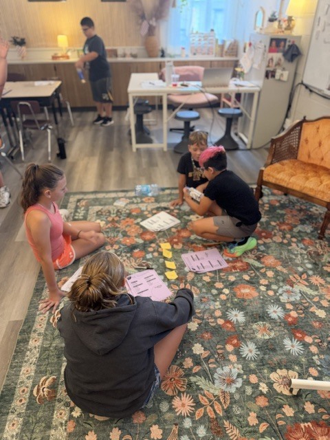 Four people sit cross-legged on a floral rug, engaged in a card game. Behind them, another person stands, and others sit at tables.