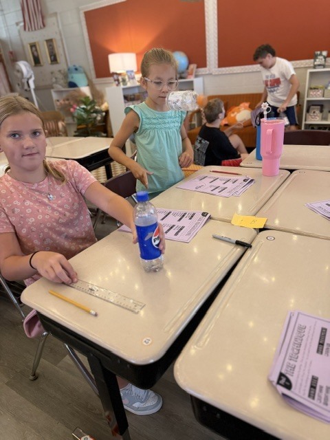 Two children at desks in a classroom, one with a ruler, pencil, and bottle, other with paper.