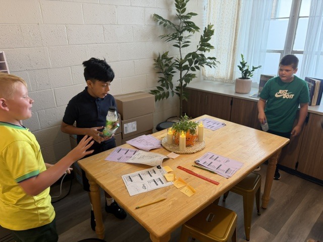 Three children are gathered around a table with papers and a candle. One holds a bottle.