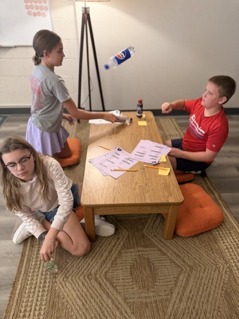 Three kids are seated on the floor, engaging in an activity with a bottle in the air.