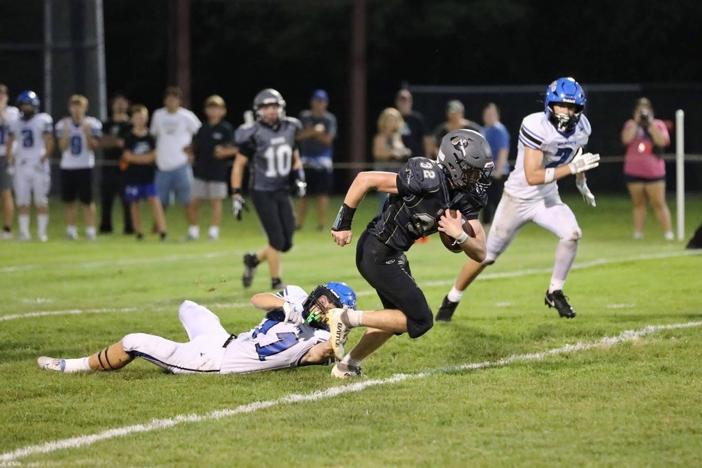 Football players run on a field. One player attempts to tackle another. People watch from the sidelines.