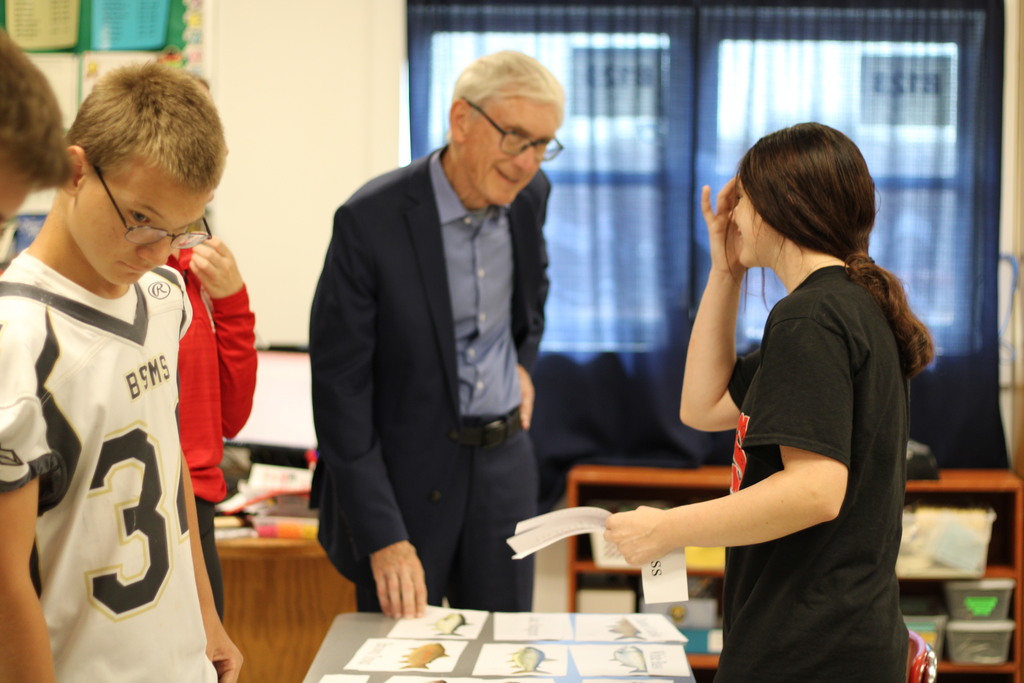 A man in a suit speaks with two students at a table with papers and cards in a classroom.