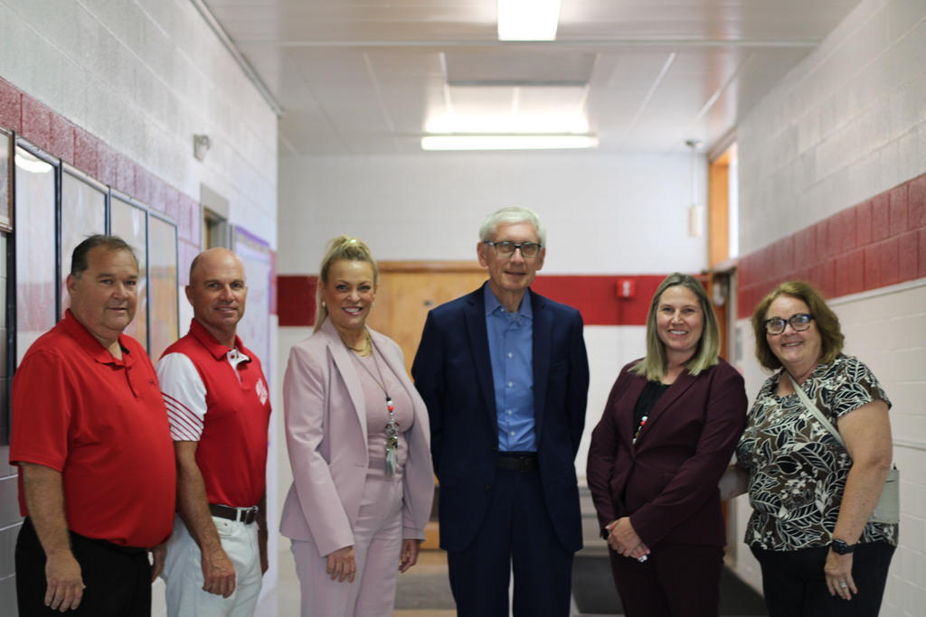 A group of six adults stand in a school hallway with red and white walls. They are all smiling.
