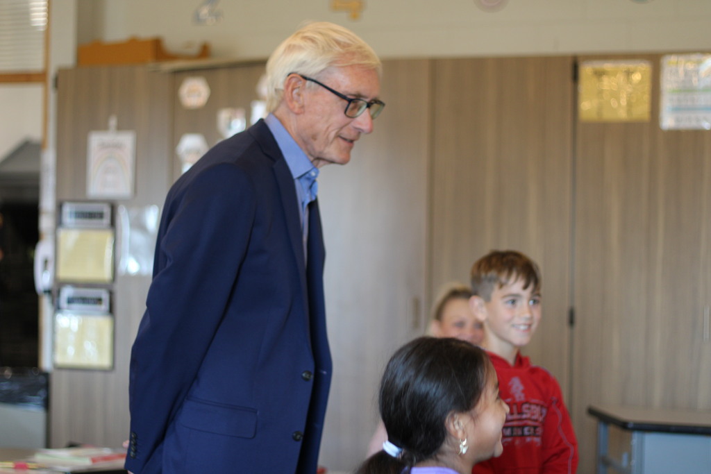 Older man in a blue suit and glasses, standing and smiling in a classroom with kids.