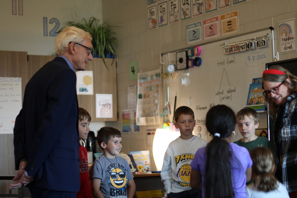 A man in a suit speaks to a group of children standing in a classroom with a whiteboard and decorations.