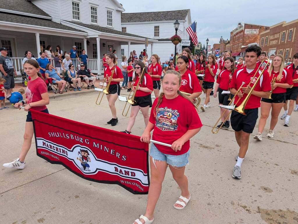 A marching band in red uniforms holds a banner while walking in a parade. Behind them are spectators and buildings.