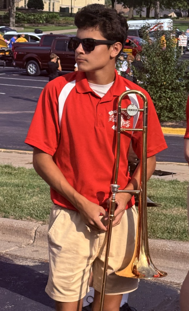 Person wearing red shirt and khaki shorts holding a trombone on a sunny day.