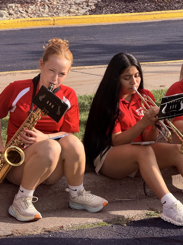 Two women in red shirts playing instruments. One holds a saxophone, the other a trumpet. They sit on the sidewalk with a curb behind them.