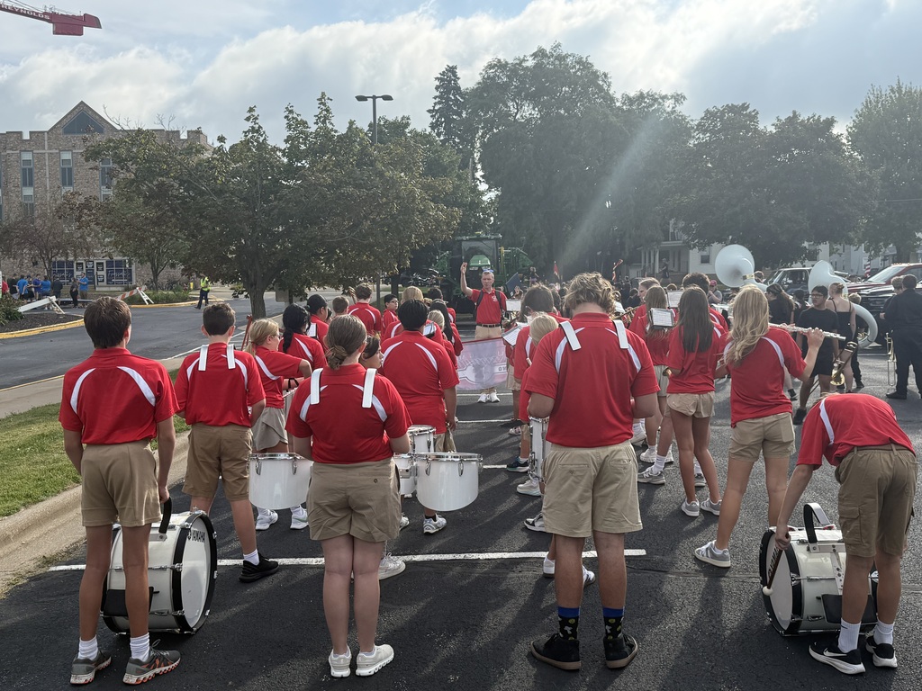 A marching band in red shirts and shorts practices on a street with trees and buildings.