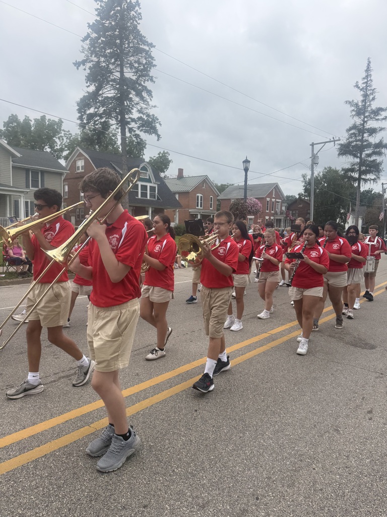 A marching band in red shirts and khaki shorts plays instruments while walking down a road.