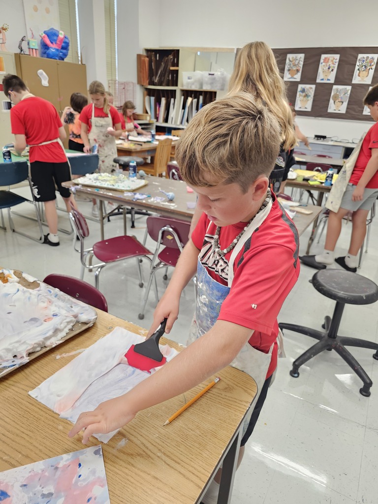 A young boy wearing a red shirt and an apron is using a squeegee to spread paint on paper.