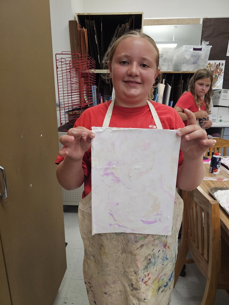 A young girl with a white apron holds up a piece of paper with a purple drawing.