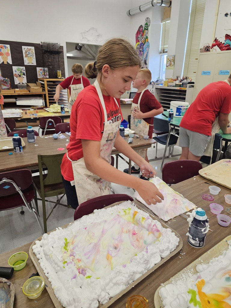 A girl in a red shirt and apron paints a colorful picture on a table in an art room.