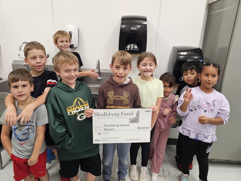 Children gather for a photo with a check. One boy holds a large check. Behind them is a sink with dispensers.