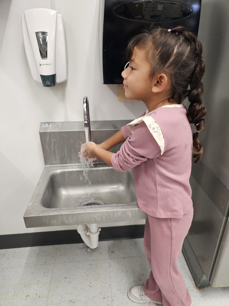 A young child washing her hands at a sink, using soap from a dispenser above.