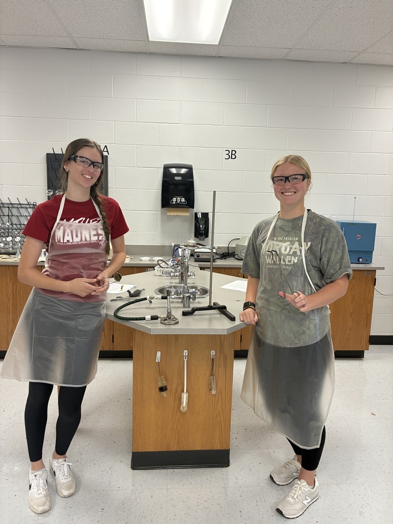 Two women in safety gear stand next to a lab station with equipment. One wears a red shirt, and the other a gray one. They both have glasses.