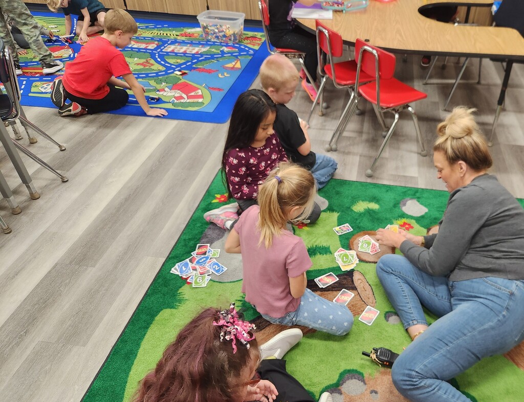 A group of children and an adult woman sit on a rug in a classroom, playing cards.