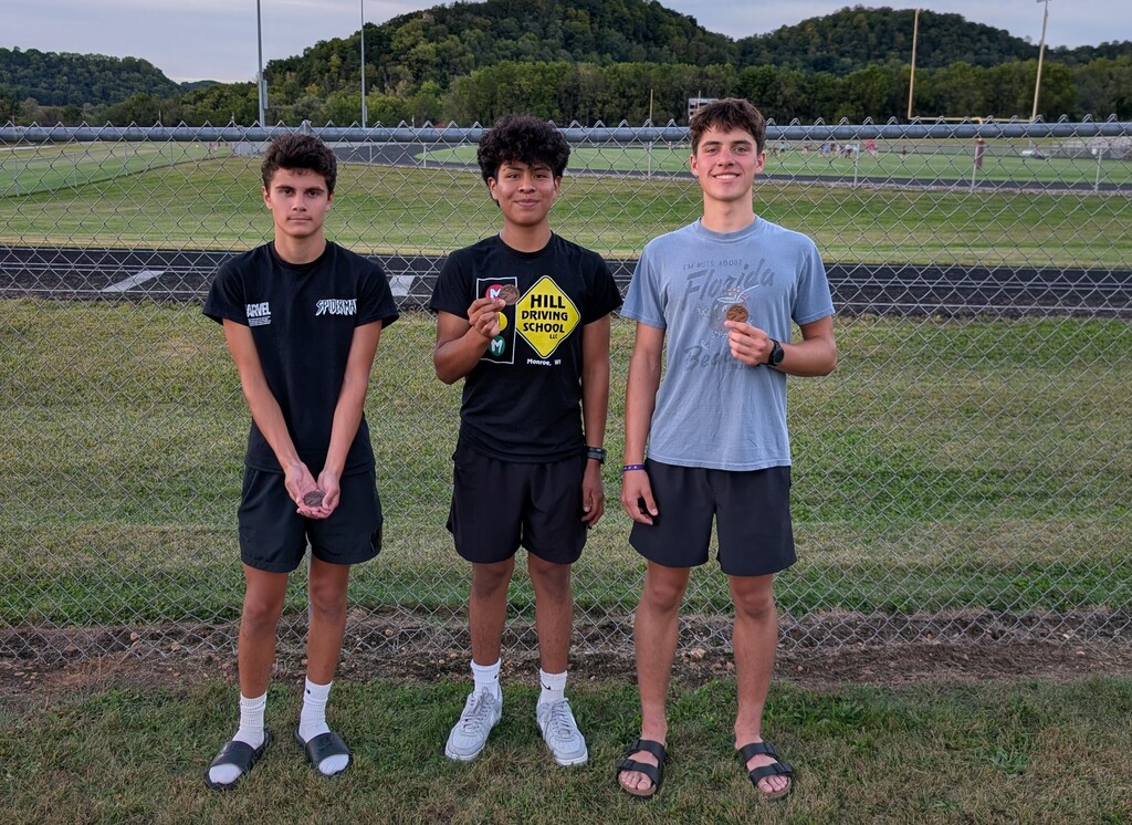 Three people stand in a grassy area near a fence with a track in the background.