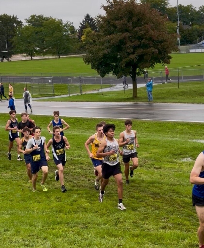 Runners in sneakers compete on a grassy field with a wet track, trees, and a fence.