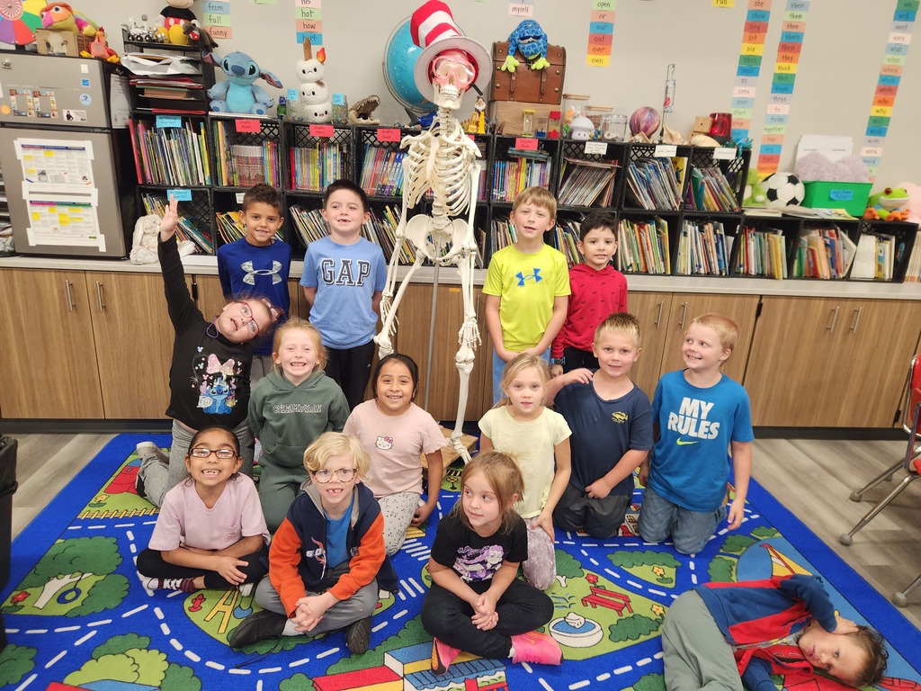Children in a classroom crouch or sit around a skeleton, with a colorful rug and bookshelves in the background.
